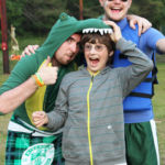 A camper dressed up in a green alligator costume and hats posing with another camper and counselor. It's for one of the themed special events on the camp program schedule